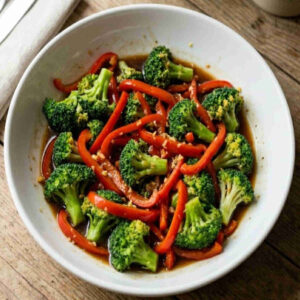 A top-down photograph of a vibrant broccoli and red pepper stir-fry presented in a white ceramic bowl. The dish features a mix of bright green broccoli florets and thinly sliced red bell peppers coated in a glossy, savory soy-ginger glaze. Small flecks of minced garlic and grated ginger are visible on the vegetables. The bowl rests on a rustic wooden table, with a neutral-colored linen napkin and the edge of a spoon visible on the upper left. The natural lighting highlights the fresh, tender-crisp textures of the food.
