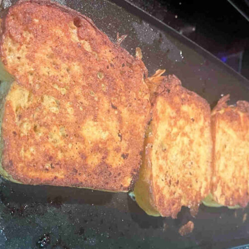 Four thick-cut slices of golden brown French toast cooking on a large electric griddle, showing the rich texture of homemade bread machine loaf.