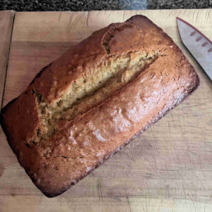 A golden-brown loaf of heritage peanut butter banana bread on a rustic wooden cutting board, featuring a professional-grade score line down the center and a dense, satiating texture.